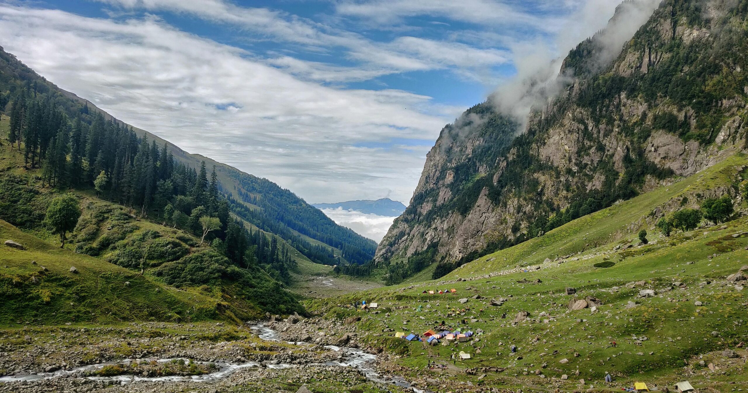 Individuals & Families Scenic view of a peaceful camping site in a lush Himalayan valley with tents and a flowing river.